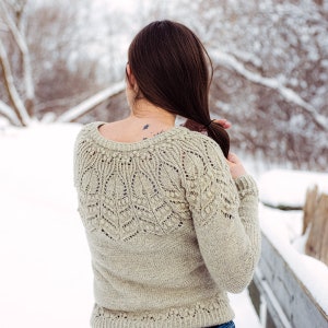 May include: A woman wearing a beige knitted sweater with a delicate lace pattern. The sweater has a round neckline and long sleeves. The woman is standing in a snowy outdoor setting.