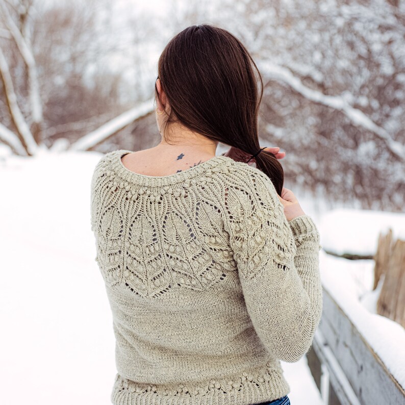 May include: A woman wearing a beige knitted sweater with a delicate lace pattern. The sweater has a round neckline and long sleeves. The woman is standing in a snowy outdoor setting.