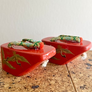 Puede incluir: Un par de sandalias geta japonesas tradicionales. Los zuecos de madera están pintados de rojo vibrante con diseños de grullas doradas. Las correas son verdes y doradas, y la base tiene una cuerda de algodón blanca.