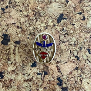 May include: An oval-shaped gold-tone metal pin featuring a blue eagle emblem, a red accent, and a small red gemstone at the top. The text reads CONTINENTAL AIR LINES with the number 5 at the bottom, set against a vertical striped background.