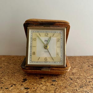 Op de afbeelding: Een vintage reiswekker met een vierkante wijzerplaat en een goudkleurig metalen frame. De wijzerplaat is crèmekleurig met zwarte cijfers en wijzers. De klok is gehuisvest in een bruine leren etui. Het woord "Endura" staat op de wijzerplaat.