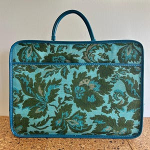 Könnte beinhalten: Vintage Handtasche mit teal-grünem Blumenmuster, passendem Griff und Besatz. Die rechteckige Tasche hat eine Vordertasche und einen oberen Griff. Das Blumenmuster kombiniert Türkis, Grün und Grau.