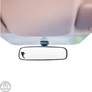 May include: A black silhouette of a fairy is on the rearview mirror of a car. The mirror is white and has a black frame.