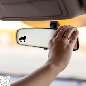 May include: A black silhouette of a dog is affixed to a car's rearview mirror. The mirror is being adjusted by a hand wearing a silver ring. The background is blurred, suggesting motion or a sunny outdoor setting.