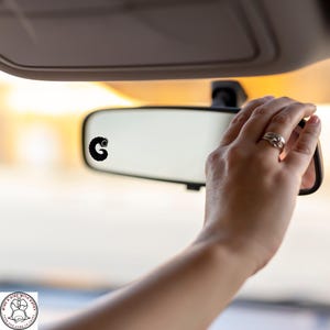 May include: A black caterpillar sticker is affixed to a car's rearview mirror. The image shows a hand adjusting the mirror, with the sticker in focus. The background is blurred, suggesting a sunny outdoor setting.