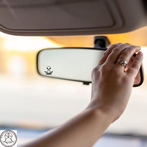 May include: A car's rearview mirror with a small black alien sticker in the center. A hand with rings is adjusting the mirror. The background is blurred, suggesting a sunny outdoor setting.