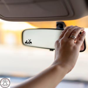 May include: A car's rearview mirror with a black silhouette of a deer family. The mirror is being adjusted by a hand with a ring. The background is blurred, suggesting a sunny day.