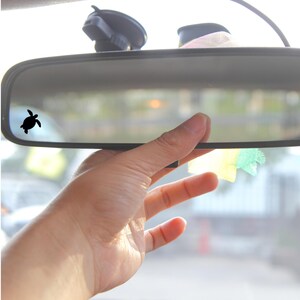 May include: A black rearview mirror with a small black turtle sticker on the left side of the mirror. A hand is reaching up to touch the mirror.