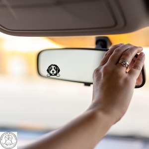 May include: A black and white dog decal is affixed to a car's rearview mirror. The decal features a stylized illustration of a dog's face, peeking over the edge of the mirror. The background is blurred, suggesting the car is in motion.