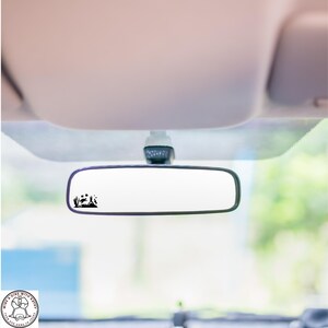 May include: A black car rearview mirror with a small black graphic of a landscape scene. The scene includes a mountain, trees, and a deer. The mirror is mounted inside a car.
