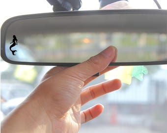 May include: A black silhouette of a mermaid is visible on the rearview mirror of a car. The mirror is attached to the windshield of the car.
