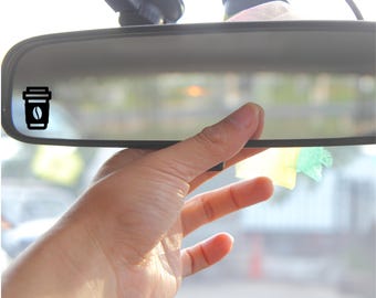 May include: A black and white car mirror sticker of a coffee cup with a coffee bean inside. The sticker is on the rearview mirror of a car.