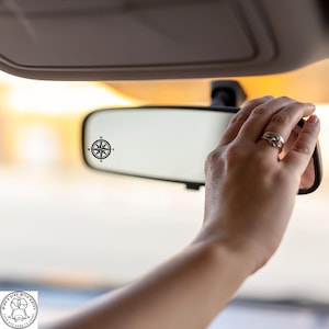 May include: A black compass rose decal is affixed to a car's rearview mirror. The mirror is being adjusted by a hand with rings. The background is blurred, suggesting a sunny outdoor setting. The compass rose design includes the cardinal directions.