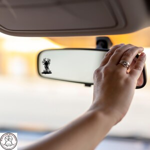May include: A black silhouette of a figure with tree-like branches is reflected in a car's rearview mirror. The mirror is being adjusted by a hand with rings. The background is blurred with warm tones.