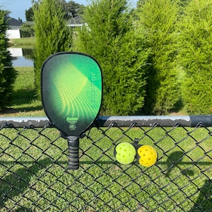 May include: A green pickleball paddle with the word "JOURNEY" printed on it, hanging on a black chain link fence. Two yellow pickleball balls are attached to the fence with a black clip.