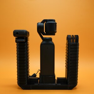 May include: A black camera stabilizer with a built-in microphone and a black tripod stand. The stabilizer is mounted on the tripod stand.