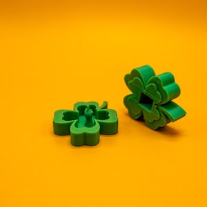 May include: Two green four-leaf clover shapes, one with a small green figure inside, on a solid orange background.