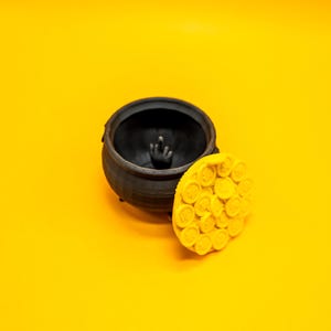 May include: A black cauldron with a lid shaped like a honeycomb. The lid is open and the cauldron is empty. The cauldron is on a yellow surface.