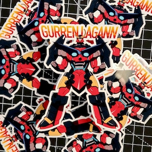 Könnte beinhalten: Eine Ansammlung von Aufklebern mit einem roten, schwarzen und gelben Roboter mit dem Text "GURREN LAGANN". Der Roboter hat ein detailliertes Design mit blauen Akzenten und befindet sich in einer dynamischen Pose. Die Aufkleber sind auf einer schwarzen Rasteroberfläche verstreut.