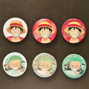 May include: Six round pinback buttons featuring anime characters. Three buttons depict a smiling character with a straw hat and red shirt. The other three buttons show a green-haired character with a sword.