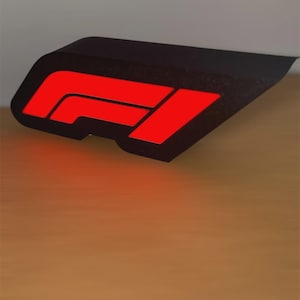 Pode incluir: Uma placa preta e angular com o logotipo vermelho da Fórmula 1. A placa tem uma superfície texturizada e está posicionada sobre uma superfície de madeira. O logotipo vermelho é iluminado, criando um contraste marcante com o fundo preto.