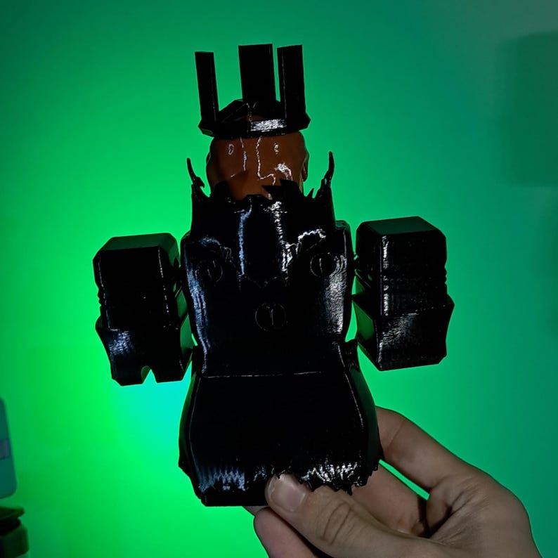 Puede incluir: Una figura negra impresa en 3D de un personaje que lleva una corona y una armadura. La figura tiene una cabeza marr&oacute;n y est&aacute; sostenida en la mano de una persona sobre un fondo verde. La figura tiene un dise&ntilde;o en bloques.