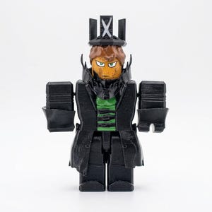 Puede incluir: Figura impresa en 3D de un personaje con abrigo negro, sombrero y rasgos cuadrados. La figura tiene una cara bronceada con ojos verdes y cabello casta&ntilde;o. Se ve una camisa verde debajo del abrigo. El sombrero tiene un dise&ntilde;o de cruz.