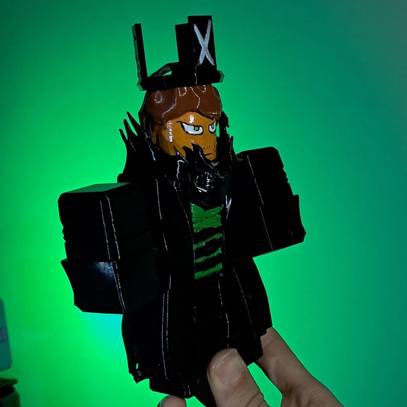 Puede incluir: Figura impresa en 3D de un personaje con abrigo negro, sombrero de copa con X y camisa a rayas verdes y negras. La figura tiene cabello casta&ntilde;o, rostro naranja y barba negra. El fondo es un degradado verde.