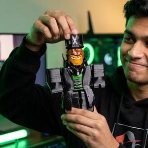 Puede incluir: Una figura impresa en 3D de un personaje con un abrigo negro, una camisa verde y un sombrero de copa con una X. La figura tiene una cara naranja con ojos amarillos y cabello casta&ntilde;o. La figura es sostenida por una persona.