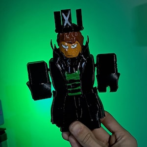 Puede incluir: Una figura impresa en 3D de un personaje con un atuendo negro, una corona con un s&iacute;mbolo "X" y una franja verde en el pecho. La figura tiene cabello casta&ntilde;o y una cara amarilla con ojos verdes. Fondo verde.