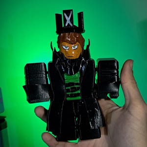 Puede incluir: Una figura impresa en 3D de un personaje con un abrigo negro, una camisa verde y una corona con una "X". La figura tiene una cara bronceada con ojos verdes y cabello casta&ntilde;o. El fondo es un degradado de verde.