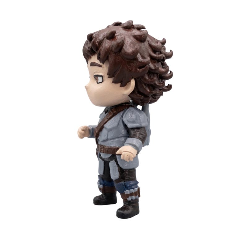 May include: A small figurine of a character with brown curly hair, wearing a gray tunic, brown belt, and dark blue pants. The figure has a small backpack and black boots. The figurine is standing on a white surface.
