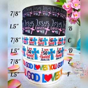 May include: A stack of six decorative ribbons with religious phrases. The ribbons are black, white, and blue, with text such as "Jesus Loves Me," "I am so loved," and "God Loves You." The ribbons vary in width, with measurements of 2.2 cm and 3.8 cm.