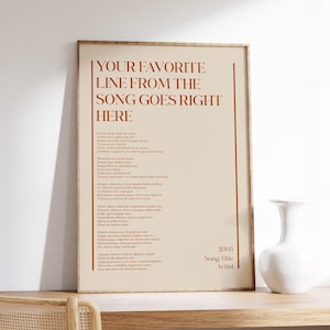 May include: A framed poster with the text "YOUR FAVORITE LINE FROM THE SONG GOES RIGHT HERE" in a serif font. The poster has a beige background with a poem and the text "2005 Song Title Artist". A white vase sits on a wooden surface.