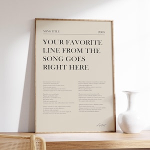 Op de afbeelding: Een ingelijste poster met de tekst "YOUR FAVORITE LINE FROM THE SONG GOES RIGHT HERE" in een minimalistisch ontwerp. De poster zit in een houten lijst en wordt op een houten oppervlak weergegeven. Rechts staat een witte vaas.