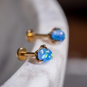 May include: A pair of gold-colored stud earrings with a round, blue opal stone. The opal has a slight iridescent quality. The earrings are set in a claw-like setting. The earrings are displayed on a white marble surface.