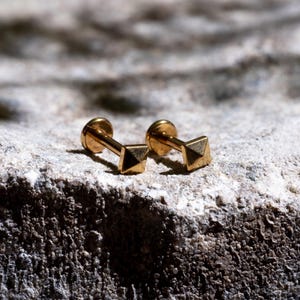 May include: A pair of gold-coloured stud earrings, each with a pyramid-shaped design. The earrings are set on a textured, light-coloured surface. The studs have a post and a flat back.
