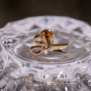 Könnte beinhalten: Ein goldfarbener Ohrstecker in Form einer Schere, mit einem kleinen Diamanten am Drehpunkt. Der Ohrring befindet sich auf einer transparenten, strukturierten Glasoberfläche.