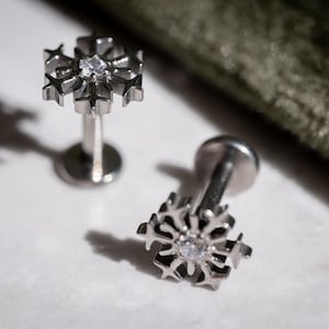 May include: Two silver-colored snowflake-shaped lip piercing studs with a clear gem in the center. The studs are on a white surface with a dark green textured background. The studs have a flat back and a post for insertion.