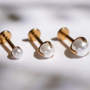 Op de afbeelding: Drie goudkleurige oorstekers met witte parels. De oorbellen zijn in een rij gerangschikt, waarbij de parelgrootte van links naar rechts toeneemt. De parels zijn rond en hebben een glanzende afwerking. De oorbellen hebben een pin en een platte achterkant.