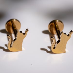 以下が含まれることがあります： 金色の猫の形をしたスタッドピアス2つ。各イヤリングは、小さな丸いポストと猫のシルエットが特徴です。猫は尻尾を丸め、足を上げてポーズをとっています。イヤリングは白い表面に表示されています。