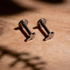 May include: Two silver-colored lip piercing studs with a cross design. The studs have a flat, round top and a post with a cross-shaped end. They are displayed on a light brown wooden surface.