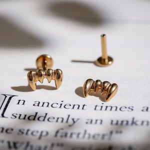 May include: Gold-coloured vampire fang earrings and a post earring are displayed on a book page with text. The earrings are in the shape of fangs, with three points each. The text on the page reads "In ancient times..."