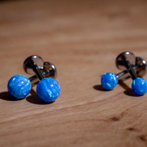 May include: Two pairs of cufflinks with a silver-tone metal base and bright blue, spherical opal-like stones. The cufflinks are displayed on a light brown wooden surface, with the light reflecting off the polished metal.