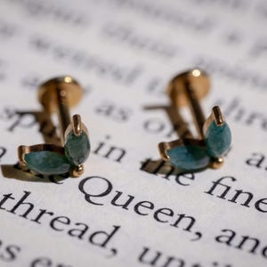 May include: A pair of gold-coloured stud earrings featuring teal-coloured teardrop-shaped stones. The earrings are set against a backdrop of printed text, with the word "Queen" visible. Jewellery.