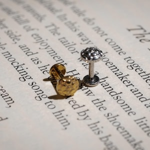 May include: Two stud earrings, one gold and one silver, rest on a book page with the text "The Two". The gold earring has a textured surface, while the silver earring has a floral design.