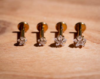 Puede incluir: Cuatro labrets dorados con detalles de cristal transparente en forma de flor. Los pendientes están dispuestos en fila sobre una superficie de madera marrón claro, con sombras creadas por la luz solar.