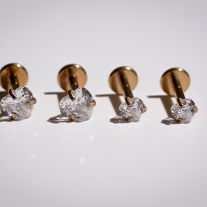 May include: Four gold-colored stud earrings with clear, heart-shaped gemstones. The earrings are arranged in a row, each casting a shadow on the white surface. The studs have a simple, elegant design.