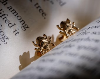 Puede incluir: Un par de pendientes dorados con forma de escarabajo. Los pendientes están colocados sobre las páginas de un libro, con el texto del libro visible en el fondo. Los pendientes tienen detalles intrincados y un acabado brillante.