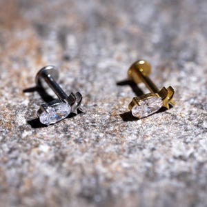May include: Two lip piercing studs. One is silver-colored, the other is gold-colored. Each stud features a clear, oval-shaped gem and a decorative star-shaped accent. The studs are displayed on a textured, gray surface.